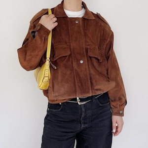 May include: A brown suede jacket with two front pockets and button fastenings. The jacket is worn with black denim jeans and a yellow shoulder bag. The jacket has a collared neckline and long sleeves. A silver belt is visible.