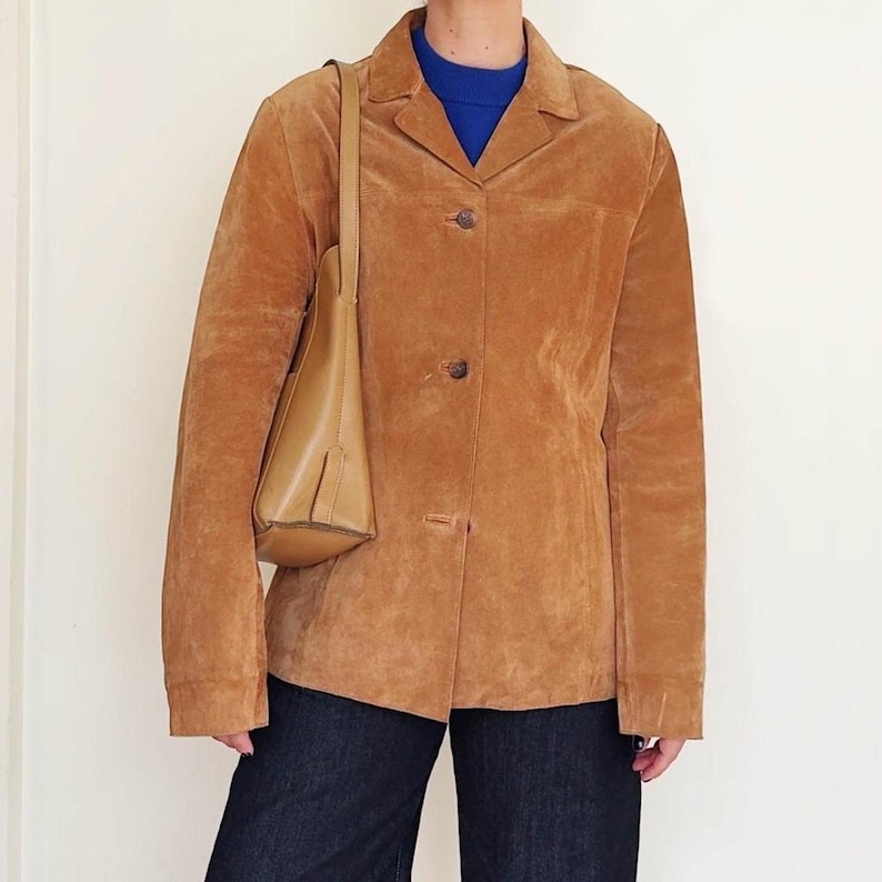 May include: A tan suede blazer with a classic collar and button closure. A matching tan leather shoulder bag is worn over the shoulder. The person is wearing a blue sweater and dark blue jeans.
