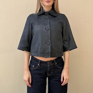 May include: Black cropped jacket with a collar and three buttons. The jacket has short sleeves and is paired with dark wash denim jeans. The outfit is a classic and versatile style.