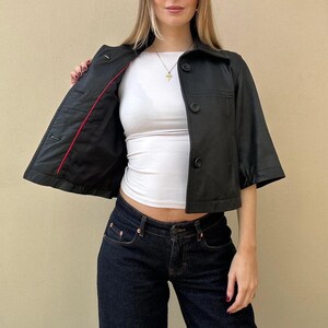 May include: A black, short-sleeved jacket with three buttons and a collar. The jacket's interior is lined with red fabric. The model is wearing a white top and dark blue jeans.