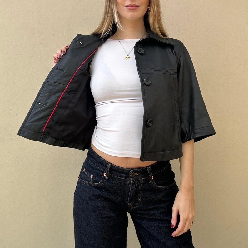 May include: A black, short-sleeved jacket with three buttons and a collar. The jacket's interior is lined with red fabric. The model is wearing a white top and dark blue jeans.