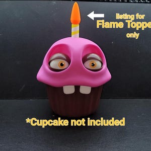 May include: A pink cupcake-shaped figurine with large, expressive eyes and two white teeth. It has a brown base and a candle with a yellow and white striped body and an orange flame. Text reads "listing for Flame Topper only" and "Cupcake not included."