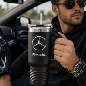 Puede incluir: Taza de viaje negra con tapa y asa plateadas. La taza presenta el logotipo de Mercedes-Benz y el nombre "Alexander" en letras plateadas. Una pajita plateada está insertada en la tapa.