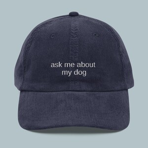 May include: A navy blue corduroy baseball cap featuring the white embroidered text "ask me about my dog". The cap has a curved peak and a rounded crown.