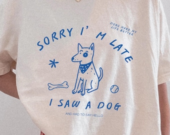 Könnte beinhalten: Cremefarbenes T-Shirt mit einer blauen Grafik eines Hundes, Knochens und Balls sowie dem Text "Sorry I'm Late, I Saw a Dog and Had to Say Hello." Das Shirt hat auch den Text "Dogs Make My Life Better."