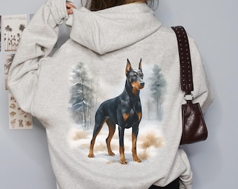 May include: A light gray hoodie featuring a Doberman Pinscher illustration on the back. The dog is black and tan, standing in a snowy forest scene. The hoodie has a drawstring hood and long sleeves.