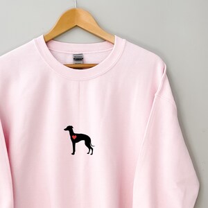 K&ouml;nnte beinhalten: Hellrosa Sweatshirt mit einer schwarzen Silhouette eines Hundes und einem roten Herz. Das Sweatshirt h&auml;ngt an einem Holzb&uuml;gel vor einem hellgrauen Hintergrund. Die Hunde-Grafik befindet sich mittig auf der Brust.