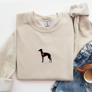 K&ouml;nnte beinhalten: Beiges Sweatshirt mit Rundhalsausschnitt, das eine schwarze Hundesilhouette mit einem roten Herz zeigt. Das Sweatshirt ist gefaltet, mit blauen Jeans und einer braunen Tasse im Hintergrund.