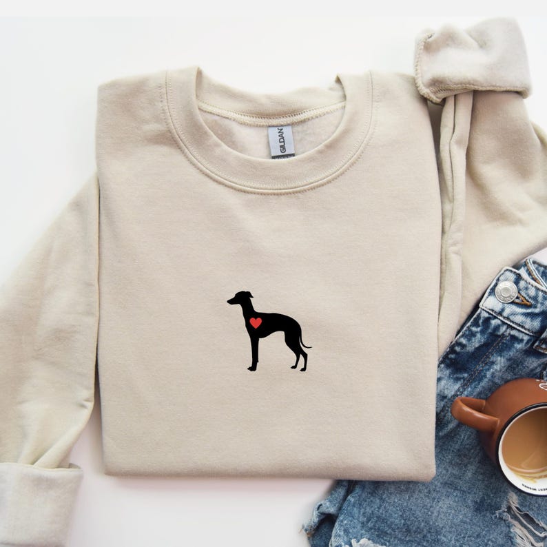 K&ouml;nnte beinhalten: Beiges Sweatshirt mit Rundhalsausschnitt, das eine schwarze Hundesilhouette mit einem roten Herz zeigt. Das Sweatshirt ist gefaltet, mit blauen Jeans und einer braunen Tasse im Hintergrund.