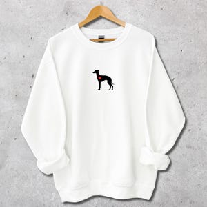 K&ouml;nnte beinhalten: Wei&szlig;es Sweatshirt mit einer schwarzen Hundesilhouette und einem roten Herz. Das Kleidungsst&uuml;ck h&auml;ngt an einem Holzb&uuml;gel. Das Design ist minimalistisch und f&uuml;r Freizeitkleidung geeignet. Der Hintergrund ist neutral.