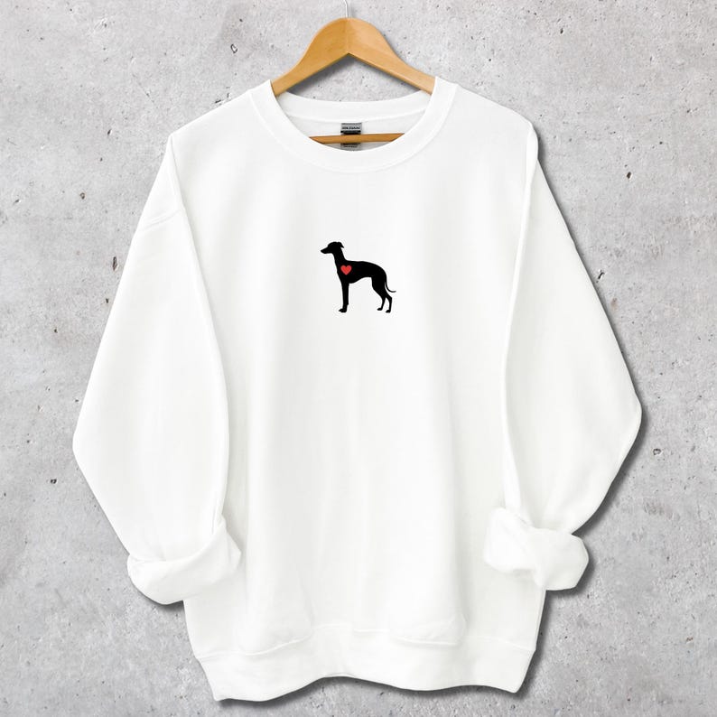 K&ouml;nnte beinhalten: Wei&szlig;es Sweatshirt mit einer schwarzen Hundesilhouette und einem roten Herz. Das Kleidungsst&uuml;ck h&auml;ngt an einem Holzb&uuml;gel. Das Design ist minimalistisch und f&uuml;r Freizeitkleidung geeignet. Der Hintergrund ist neutral.