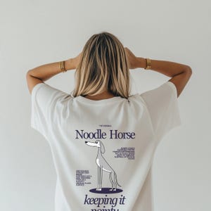 Può includere: Una maglietta bianca con la scritta "Noodle Horse" e un'illustrazione di un levriero. La maglietta contiene anche il testo "keeping it pointy" e fatti sui levrieri. Un capo d'abbigliamento casual.