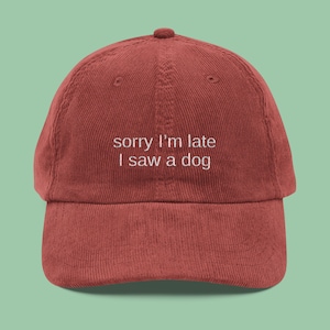 May include: A brick-red corduroy baseball cap with a curved brim. The cap features the embroidered text "sorry I'm late I saw a dog" in white, centered on the front. The cap has a rounded crown and an adjustable strap.