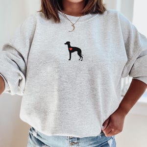 K&ouml;nnte beinhalten: Hellgraues Sweatshirt mit einer schwarzen Hunde-Silhouette und einem roten Herz. Das Sweatshirt hat lange &Auml;rmel und einen Rundhalsausschnitt. Die Hunde-Grafik befindet sich auf der Brust.