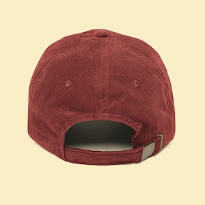 May include: A burgundy corduroy baseball cap. The cap features a curved brim, a six-panel design, and an adjustable strap with a metal buckle. The back of the cap is visible, showing the strap and the opening for the wearer's head.
