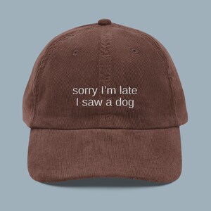May include: A brown corduroy baseball cap with the embroidered text "sorry I'm late I saw a dog" in white. The cap has a curved brim and a button on top.