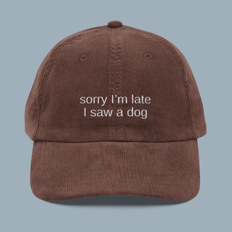 May include: A brown corduroy baseball cap with the embroidered text "sorry I'm late I saw a dog" in white. The cap has a curved brim and a button on top.
