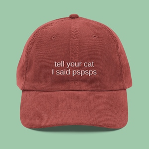 May include: A rust-colored corduroy baseball cap with a curved brim. The cap features white embroidered text that reads "tell your cat I said pspsps". The hat has a rounded crown and an adjustable strap.