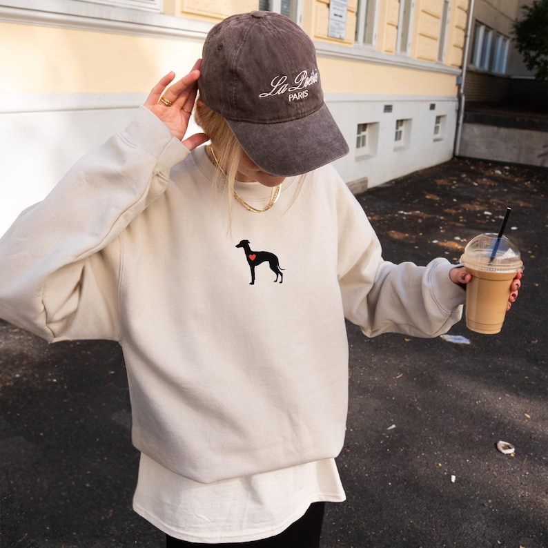 K&ouml;nnte beinhalten: Beiges Sweatshirt mit schwarzer Hunde-Silhouette und rotem Herz. Dazu eine braune Baseballkappe mit dem Text "La Poste PARIS". Das Bild zeigt auch ein Getr&auml;nk in einem transparenten Plastikbecher.