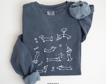 May include: A denim blue sweatshirt with white line drawings of dachshunds in various poses. Some dogs are holding balloons, bones, and flowers. The word "DENIM" is printed in white at the bottom of the sweatshirt.