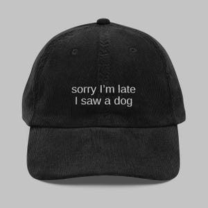 May include: Black corduroy baseball cap with the embroidered text "sorry I'm late I saw a dog" in white. The cap has a curved brim and an adjustable strap at the back.