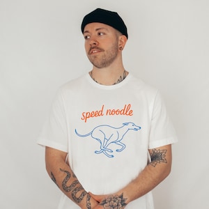 Può includere: T-shirt bianca con la scritta "speed noodle" in corsivo rosso sopra un contorno blu di un levriero in corsa. La maglietta ha maniche corte.