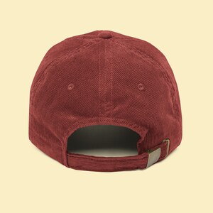 May include: A burgundy corduroy baseball cap. The cap features a curved brim, a six-panel design, and an adjustable strap with a metal buckle. The back of the cap is visible, showing the strap and the opening for the wearer's head.