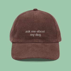 May include: A brown corduroy baseball cap with the phrase "ask me about my dog" embroidered in white. The cap has a curved brim and an adjustable strap at the back. The hat is a solid color and has a classic, casual style.