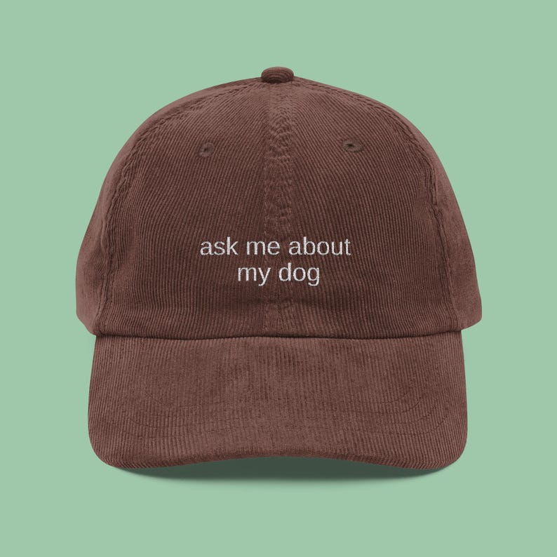 May include: A brown corduroy baseball cap with the phrase "ask me about my dog" embroidered in white. The cap has a curved brim and an adjustable strap at the back. The hat is a solid color and has a classic, casual style.