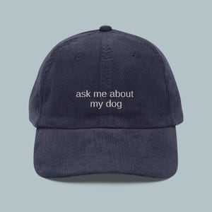 May include: Navy blue corduroy baseball cap with the text "ask me about my dog" embroidered in white. The cap has a curved brim and a rounded top.