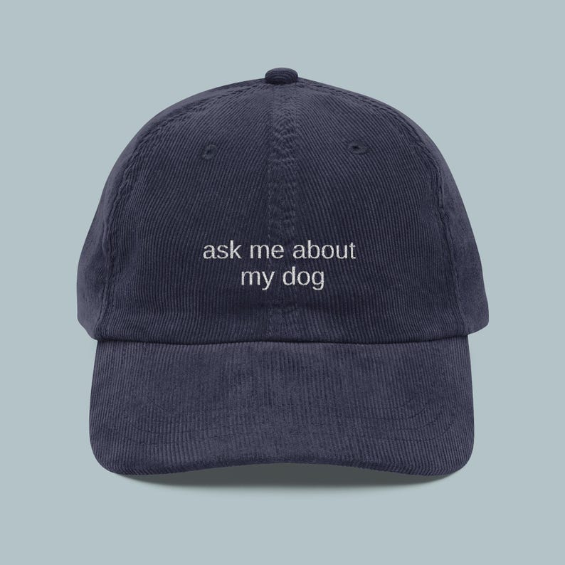 May include: Navy blue corduroy baseball cap with the text "ask me about my dog" embroidered in white. The cap has a curved brim and a rounded top.