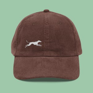 May include: A brown corduroy baseball cap with a curved brim. The cap features a white embroidered image of a running dog on the front. The cap has a six-panel design with a top button and an adjustable strap.