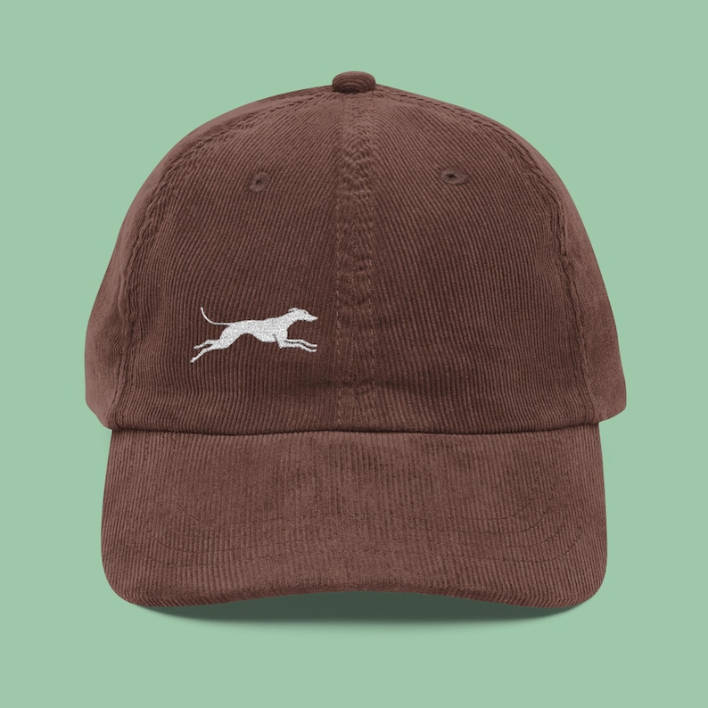 May include: A brown corduroy baseball cap with a curved brim. The cap features a white embroidered image of a running dog on the front. The cap has a six-panel design with a top button and an adjustable strap.