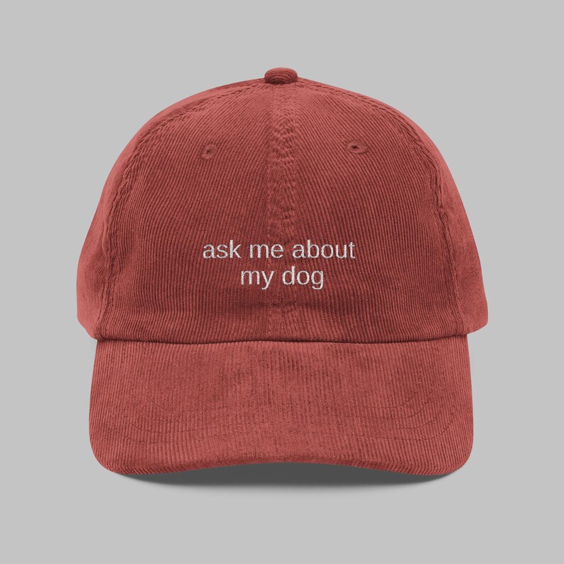 May include: A brick-red corduroy baseball cap with a curved brim. The cap features the white text "ask me about my dog" embroidered on the front. The hat has a classic six-panel design with a top button and an adjustable strap.