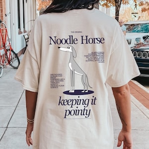 Può includere: T-shirt beige con grafica di levriero e testo "Noodle Horse" e "keeping it pointy". La maglietta include anche testo sui levrieri. Una maglietta casual e confortevole.
