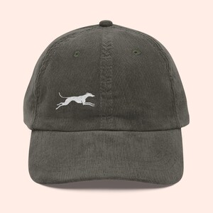 May include: Olive green corduroy baseball cap featuring a white embroidered running dog design. The cap has a curved brim and an adjustable strap at the back. A stylish accessory for casual wear.