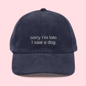 May include: Navy blue corduroy baseball cap with the embroidered text "sorry I'm late I saw a dog". The cap has a curved brim and a top button.