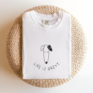 Può includere: T-shirt bianca con un disegno a tratto nero della testa di un cane e la scritta "LIFE IS GREYT" in grafia manoscritta. La maglietta è piegata e appoggiata su una superficie intrecciata marrone chiaro.