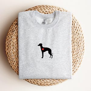K&ouml;nnte beinhalten: Hellgraues Sweatshirt mit schwarzer Hunde-Silhouette und rotem Herz. Das Sweatshirt liegt gefaltet auf einer gewebten, kreisf&ouml;rmigen Oberfl&auml;che.
