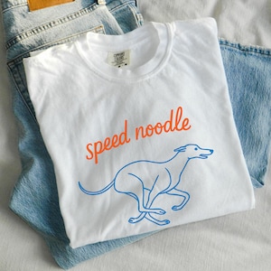 Può includere: T-shirt bianco con la scritta "speed noodle" in arancione e un contorno blu di un levriero in corsa. La maglietta è piegata sopra un paio di jeans blu chiaro.