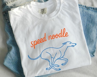 Könnte beinhalten: Weißes T-Shirt mit dem Schriftzug "speed noodle" in Orange und einer blauen Umrisszeichnung eines rennenden Windhundes. Das T-Shirt liegt gefaltet auf einer hellblauen Jeans.