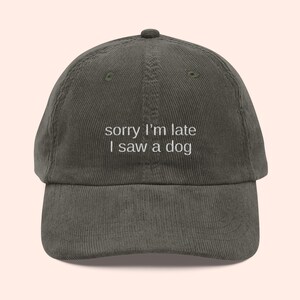 May include: Olive green corduroy baseball cap with the embroidered text "sorry I'm late I saw a dog" in white. The cap has a curved brim and a button on top. A casual accessory.