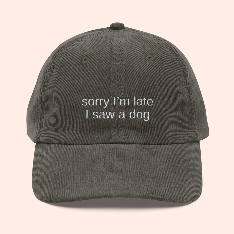 May include: Olive green corduroy baseball cap with the embroidered text "sorry I'm late I saw a dog" in white. The cap has a curved brim and a button on top. A casual accessory.