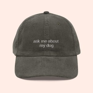 May include: Olive green corduroy baseball cap with the text "ask me about my dog" embroidered in white. The cap has a curved brim and a fabric-covered button on top.