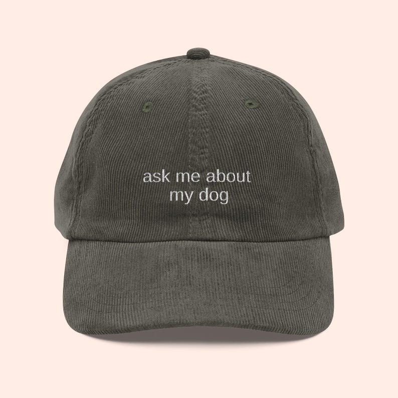 May include: Olive green corduroy baseball cap with the text "ask me about my dog" embroidered in white. The cap has a curved brim and a fabric-covered button on top.