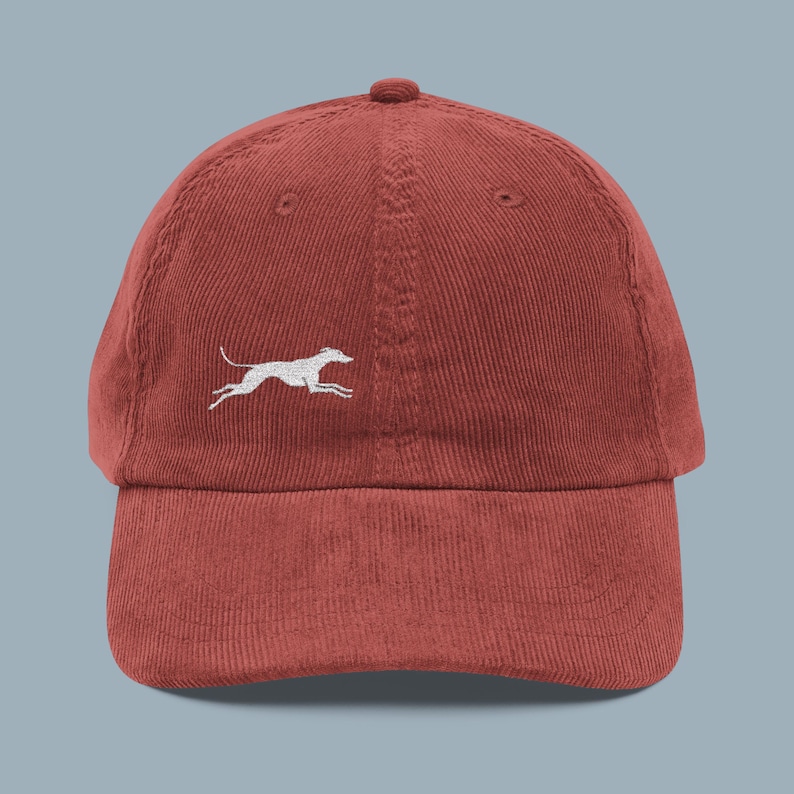 May include: A brick-red corduroy baseball cap featuring a white embroidered running dog on the side. The cap has a curved brim and an adjustable strap at the back, offering a classic, casual style. This hat is a great accessory for dog lovers.