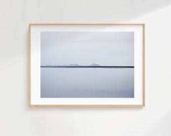 May include: A framed landscape photograph depicting a tranquil, minimalist scene. The image shows a calm body of water reflecting the overcast sky, with a distant horizon and subtle mountain silhouettes. The frame is light wood.