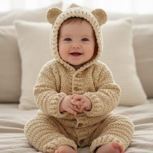 May include: A baby wearing a beige crocheted bear suit with a hood and button-up front, featuring bear ears. The outfit includes a hooded jacket and matching trousers. The baby is sitting on a bed with neutral-toned pillows in the background.