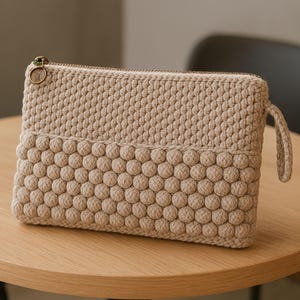 May include: A beige crochet clutch with a textured, bobble stitch design on the lower half. The rectangular bag features a gold zipper and a small loop handle. The bag is sitting on a light wood table.