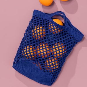 Könnte beinhalten: Eine marineblaue gehäkelte Netztasche gefüllt mit Orangen. Die Tasche hat zwei Griffe und eine rechteckige Form. Die Orangen sind leuchtend orange und durch das offene Gewebe der Tasche sichtbar. Der Hintergrund ist einfarbig rosa.